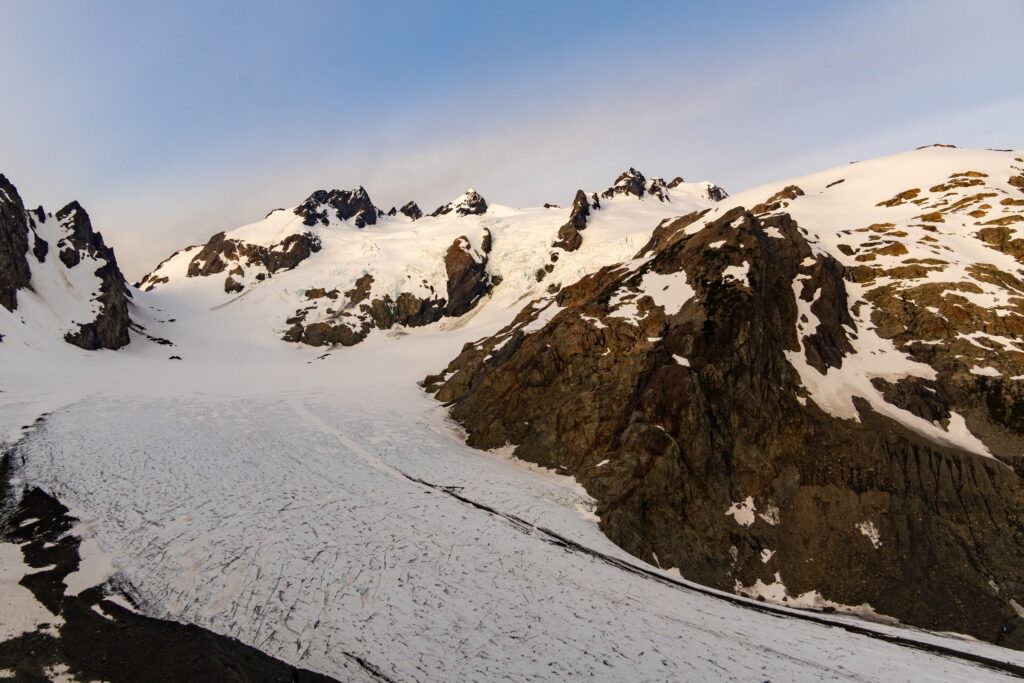 A sweeping landscape view of the vast, cracked Blue Glacier sweeping upward toward towering, snow-dusted Mount Olympus under a pale sky.