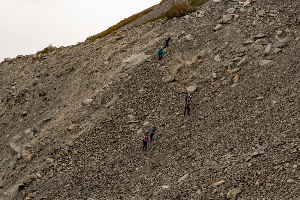 Hikers with backpacks spaced out as they carefully navigate their way up a steep, challenging slope of loose, grey scree and rock.