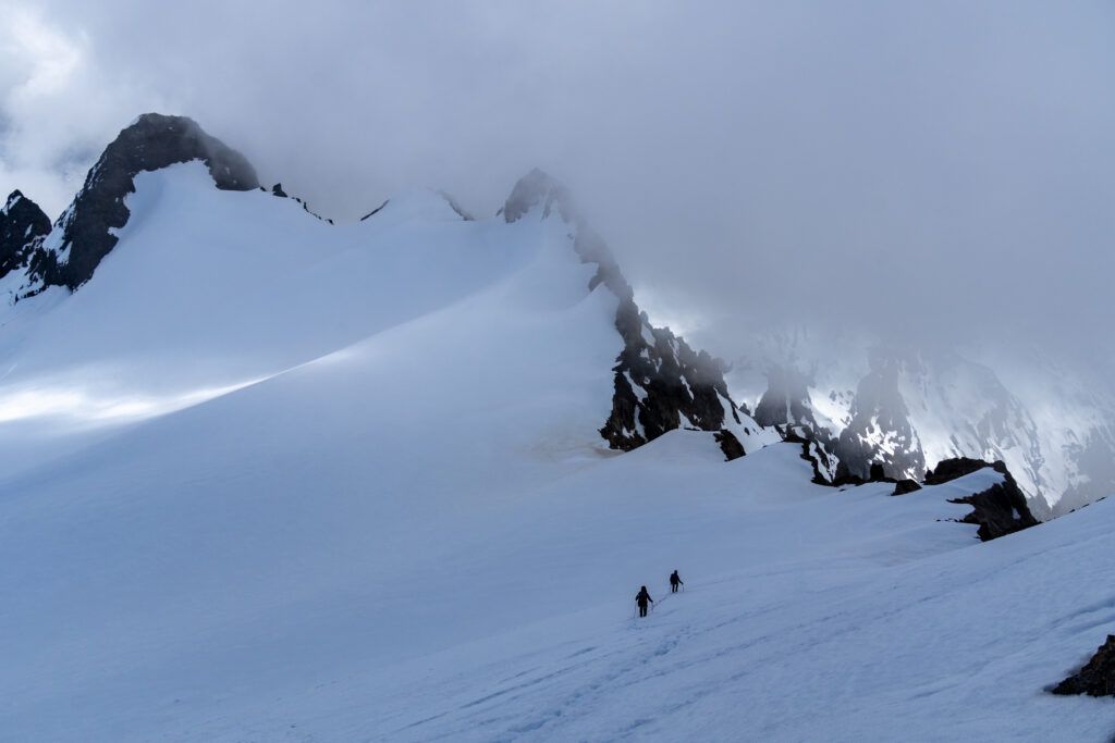Two small figures of roped mountaineers ascending a vast, snowy mountain ridge toward dark, jagged peaks that are heavily shrouded in thick, low-hanging clouds.
