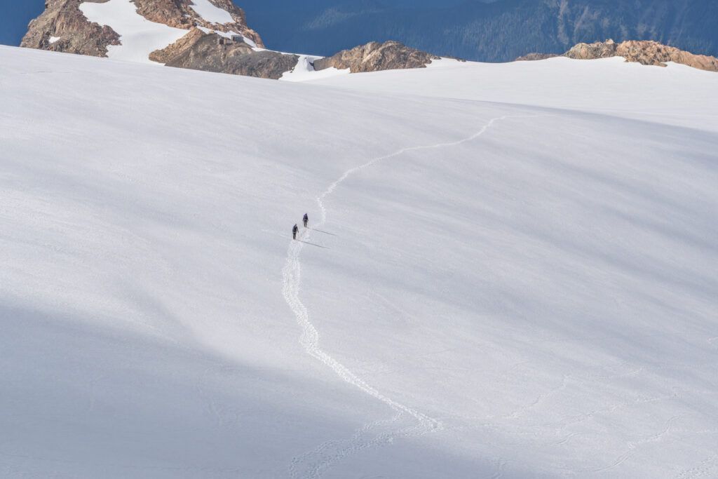 A long, winding trail of footprints left by two distant hikers walking across an enormous, smooth expanse of white snow of the Blue Glacier, with rocky ridges visible far in the background.