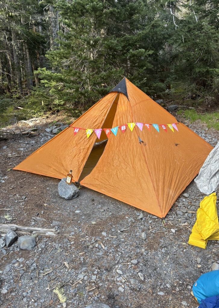 An orange pyramid-style camping tent pitched on rocky ground in a forest, decorated with a colorful "HAPPY BIRTHDAY" pennant banner strung across its partially open front doors. A small glass whiskey bottle sits on a rock right next to the tent entrance.